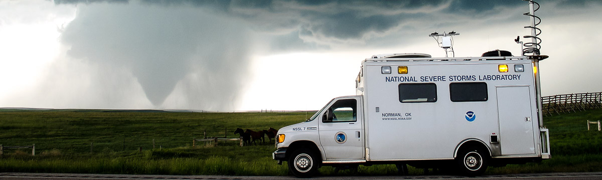 Type III ambulance-style vehicle with NOAA and NSSL logos and the words "National Severe Storms Laboratory" on the side and observation equipment mounted to the roof. The vehicle is parked at the side of a road in front of a pasture where horses are grazing. A tornado can be seen in the distance.