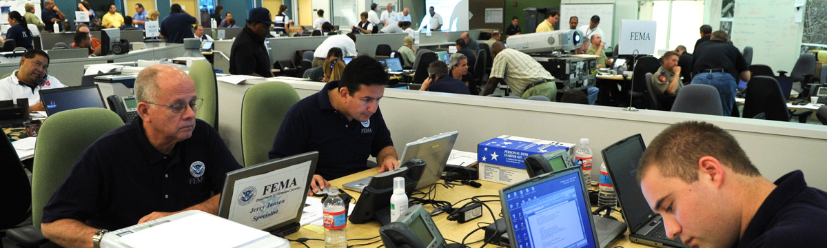 Many people at work with laptops in a large room. A sign that says "FEMA" is near the closest group, and an open laptop has a large label on the lid saying "FEMA" in the foreground.