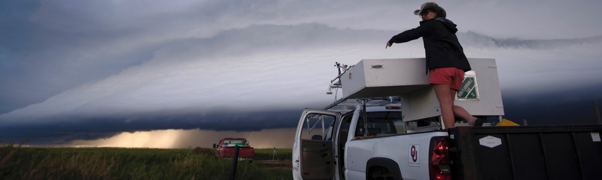 A woman rides in the bed of a white pickup truck bearing equipment in the bed. She is pointing forward. The white truck, and another red truck further down the road, are headed toward a supercell cloud.