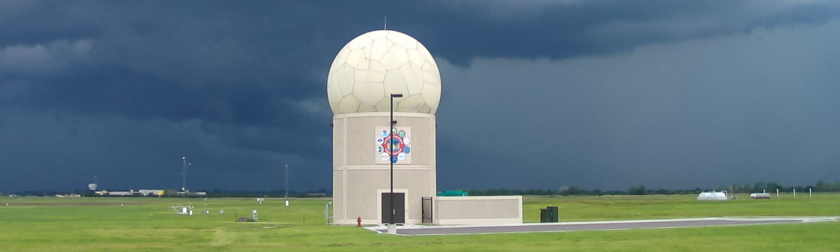 Photograph of a cylindrical building with a radome on top. The surrounding ground is covered in green grass and the sky has dark gray clouds.