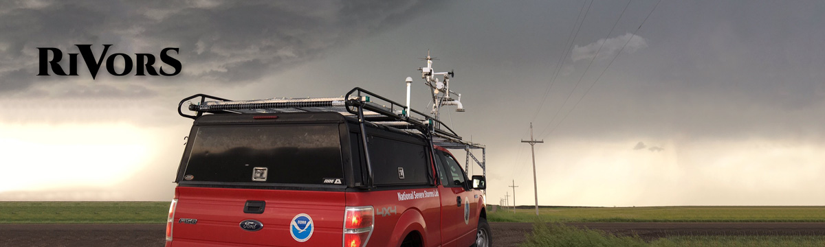 Red truck facing away from the camera, towards a storm at sunset. The truck is stopped along a country road in a rural area. It has observation equipment mounted to the top and a NOAA logo on the rear panel, along with the words "National Severe Storms Lab" and a NSSL logo on the side.