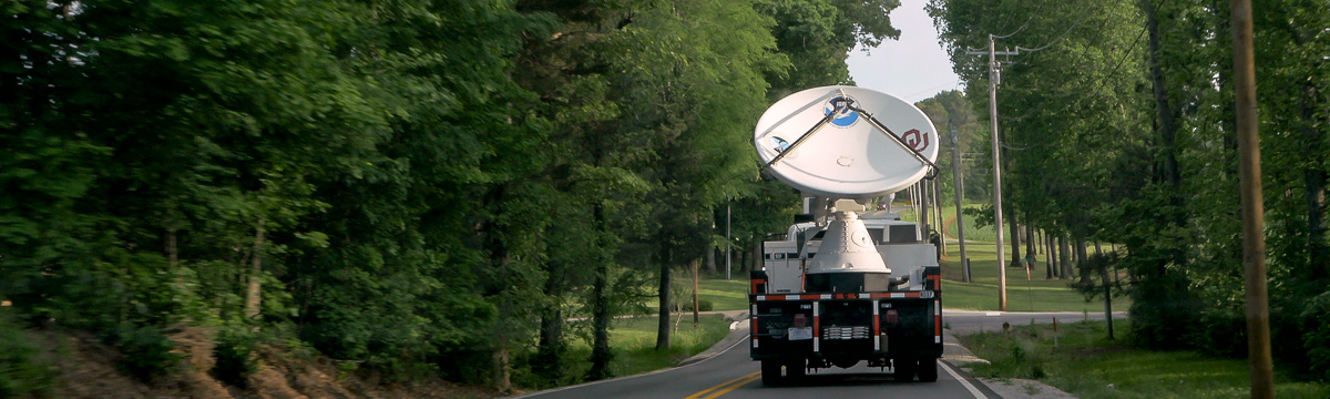 Mobile radar headed away from the camera on a two-lane road in a heavily wooded area. The radar dish has NSSL, NOAA, and University of Oklahoma logos on it.